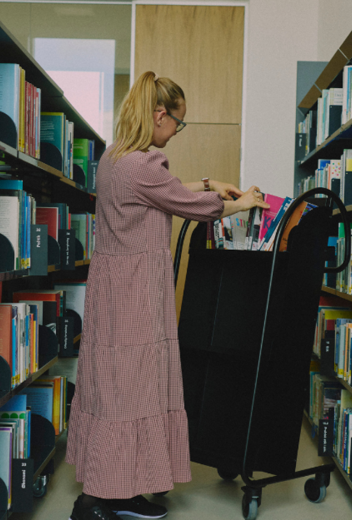 Billedet viser to bogreoler, en i venstreside og en i højreside. Reolerne er fyldt med bøger fra Absalonsbibliotek. Mellem dem går en kvindlig bibliotekar med lyst hår opsat i en hestehale. Hun er iført en lyserød gulvlang kjole og er ved at sætte bøger på plads fra en bogvogn.