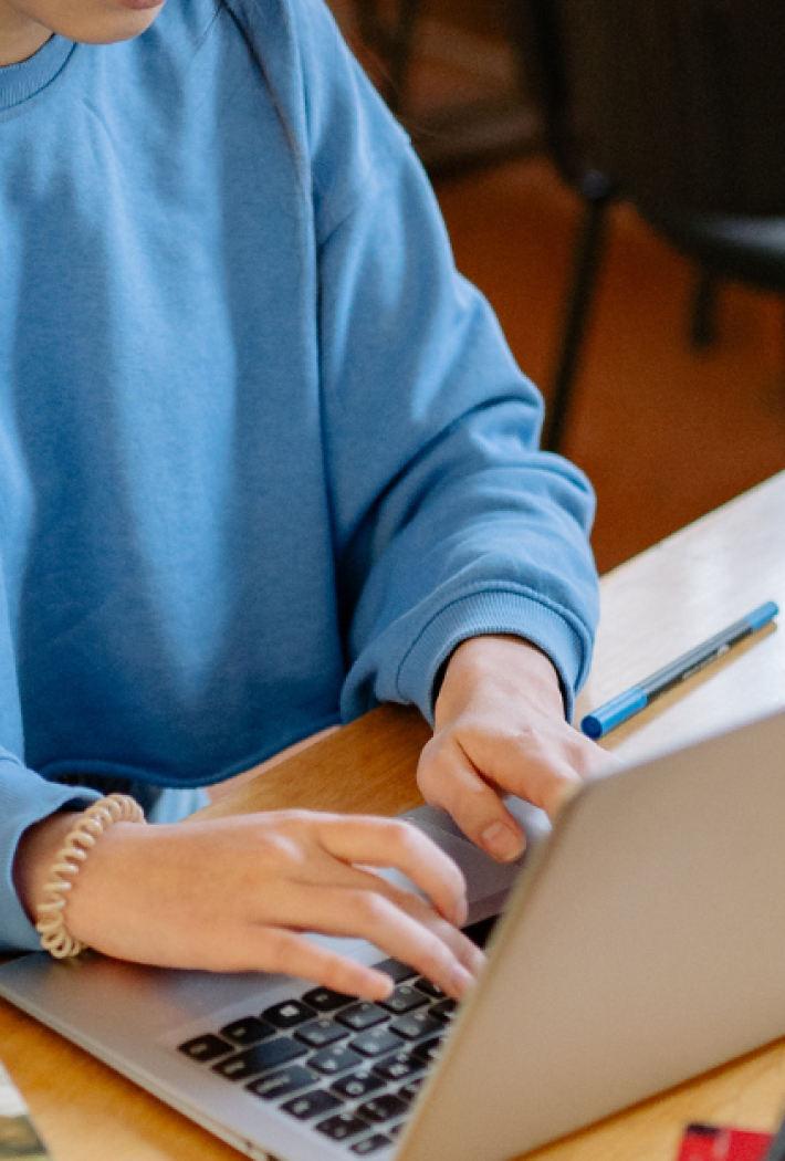 A person sits at a table in a library working on a laptop. On the table are open books, other study materials, and a pen.
